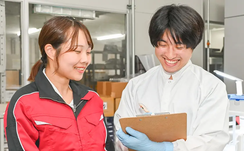 作業着の女性と白衣の男性が製造現場でクリップボードを確認しながら話し合う様子